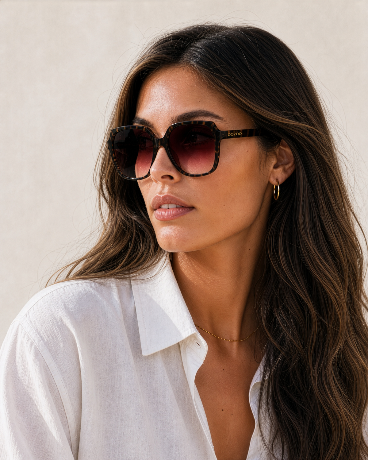 A woman with long wavy brown hair wearing oversized square sunglasses with a dark brown gradient lens and black frames, paired with a white collared shirt against a light beige background.